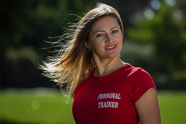 Anna outdoors in a red T-shirt reading PERSONAL TRAINER, hair blowing across her face, with a green park blurred behind her.