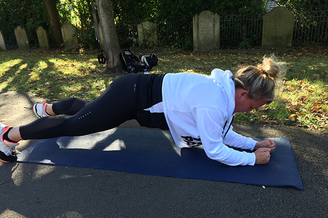 A client in a white hoodie and black leggings holding a plank on a navy mat beside a park path, face turned away.