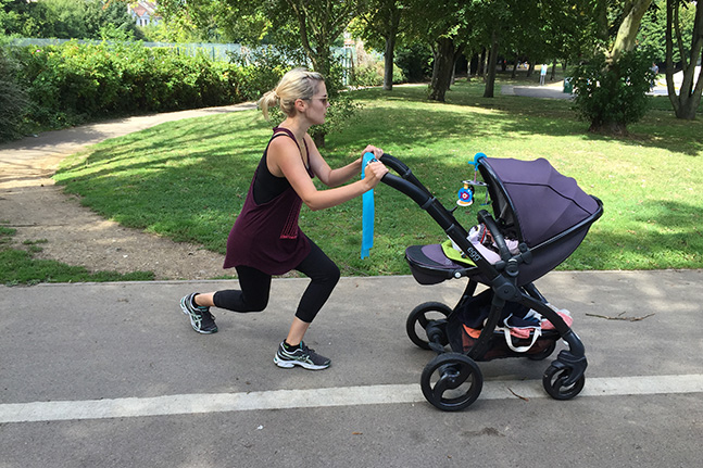 A woman in a dark vest and leggings lunging along a park path while pushing a purple buggy, face turned away from the camera.