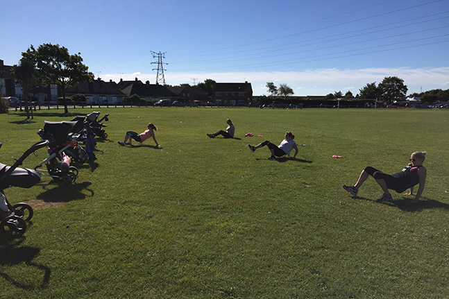A sunny recreation ground: buggies parked on the grass in the foreground and four women spread across the field doing sit-ups.